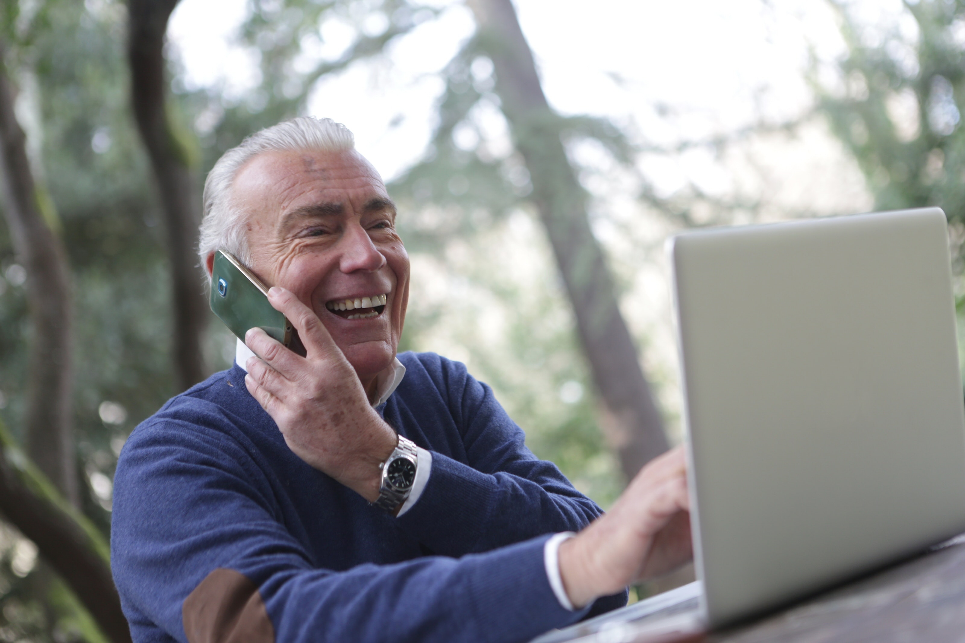 Older man outdoors on phone with laptop